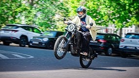 A person riding a motorcycle performing a wheelie on a street lined with parked cars.